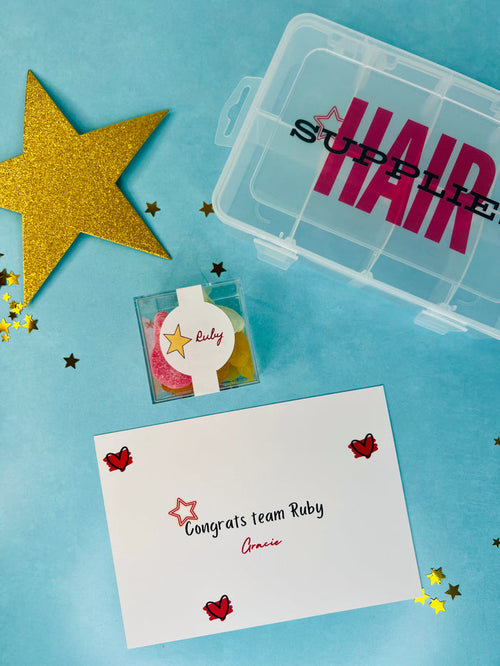 A styled flat-lay of hair supply accessories on a light blue background, including a clear plastic storage box labeled 'HAIR SUPPLIES', a small clear box with colorful hair ties, a white congratulatory card reading 'Congrats team Ruby', and a large gold glitter star.