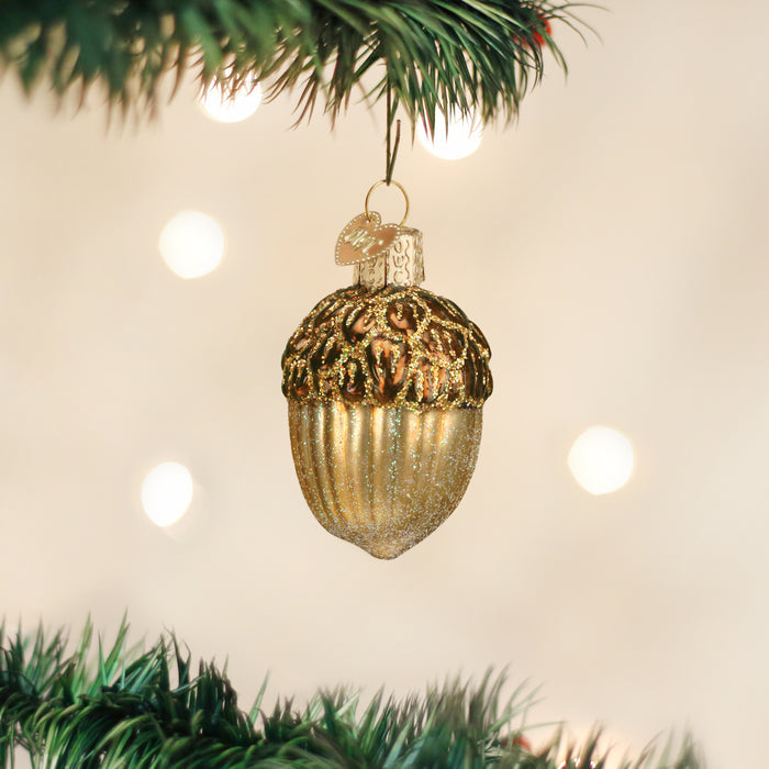A gold acorn-shaped Christmas ornament hangs from a green tree branch with blurred white lights in the background.