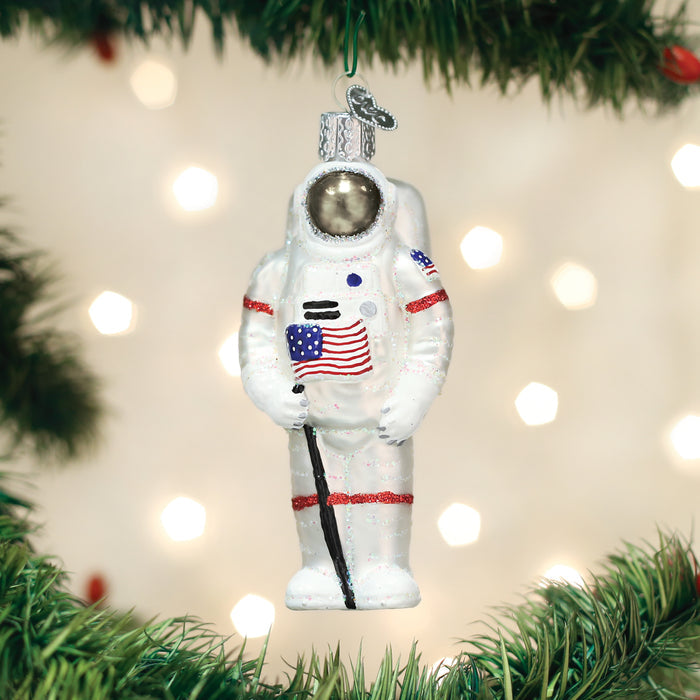 A glass Christmas ornament shaped like an astronaut holding an American flag hangs from a green tree branch, with blurred lights in the background.