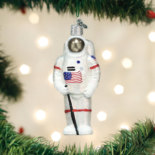 A glass Christmas ornament shaped like an astronaut holding an American flag hangs from a green tree branch, with blurred lights in the background.
