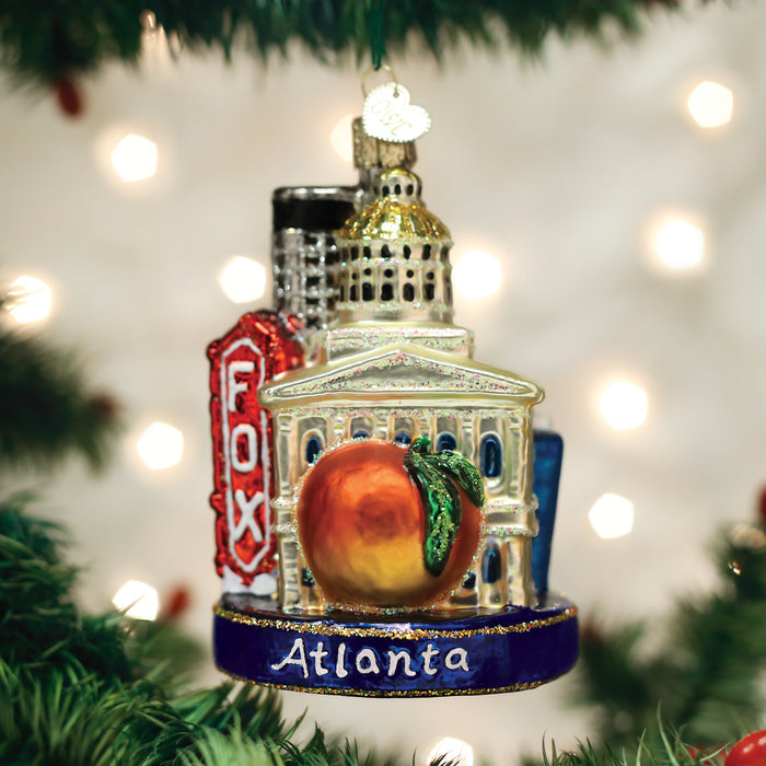 A Christmas tree ornament featuring an Atlanta cityscape, with the Capitol dome, the Fox Theatre sign, and a large peach in front, hanging on a tree with white lights in the background.