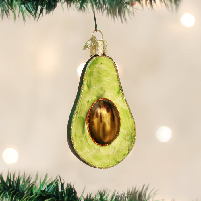 A Christmas tree ornament shaped like a half avocado with a brown pit hangs on a green branch, with a blurred background of warm lights.