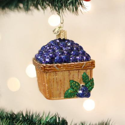 A Christmas tree ornament shaped like a wooden basket filled with glittery blue berries and decorated with green leaves, hanging from a tree branch with blurred lights in the background.