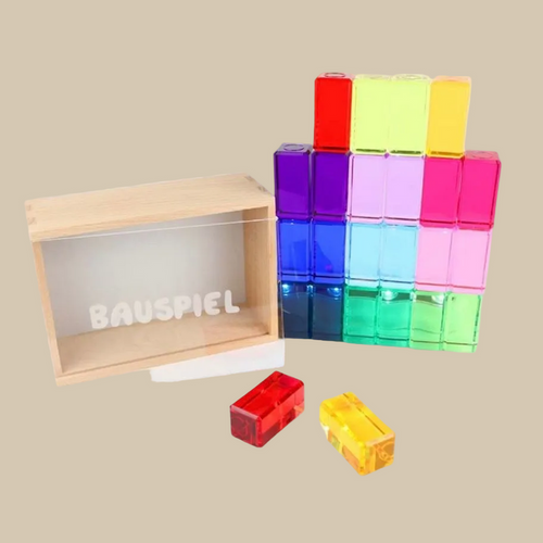 Colorful transparent building blocks with a wooden box labeled 'BAUSPIEL' on a beige background