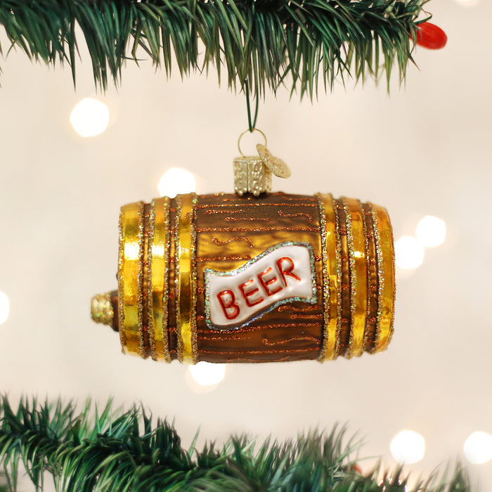 A shiny, barrel-shaped Christmas tree ornament with gold accents and the word BEER written on a white label hangs from a green pine branch against a background of blurred lights.