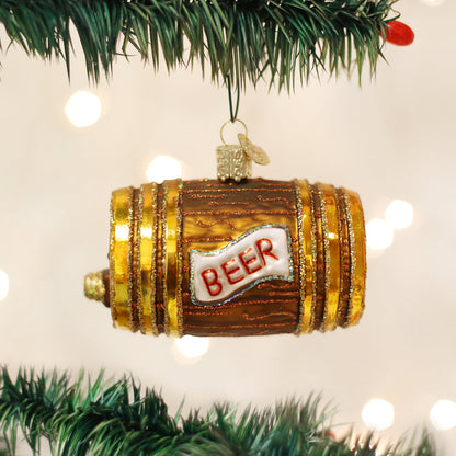 A shiny, barrel-shaped Christmas tree ornament with gold accents and the word BEER written on a white label hangs from a green pine branch against a background of blurred lights.