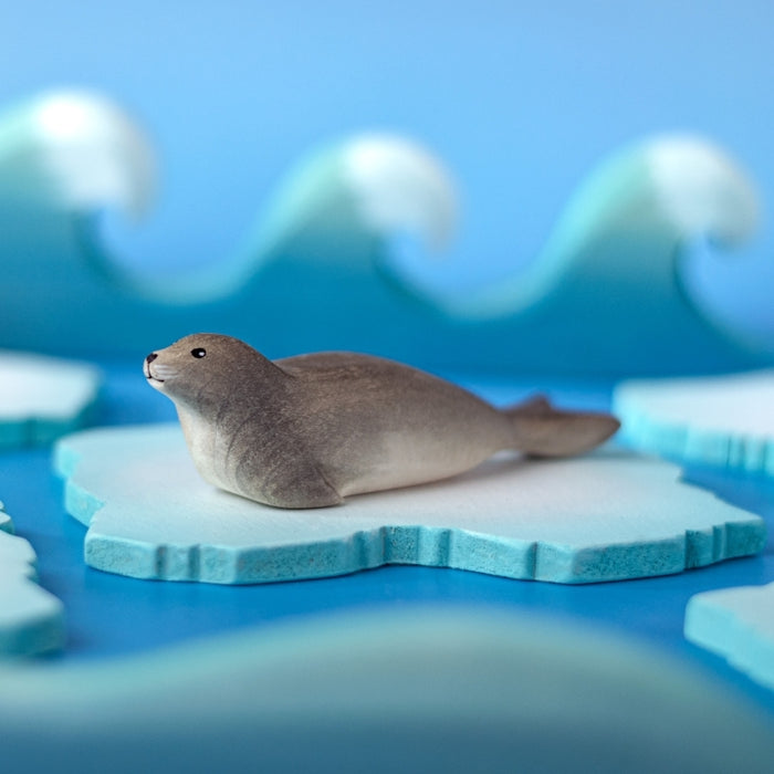 A small realistic seal figurine resting on a light blue and white iceberg, with stylized blue ocean waves blurred in the background.