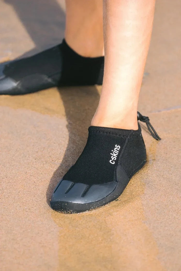 C-Skins Legend Junior Reef Boots worn on sandy beach, black neoprene with rubber sole