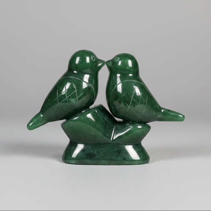 A green jade sculpture of two small birds perched on a matching base, photographed on a grey background