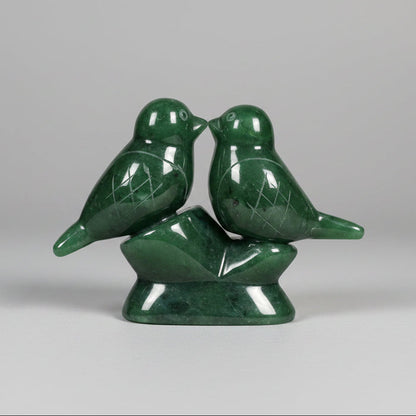 A green jade sculpture of two small birds perched on a matching base, photographed on a grey background