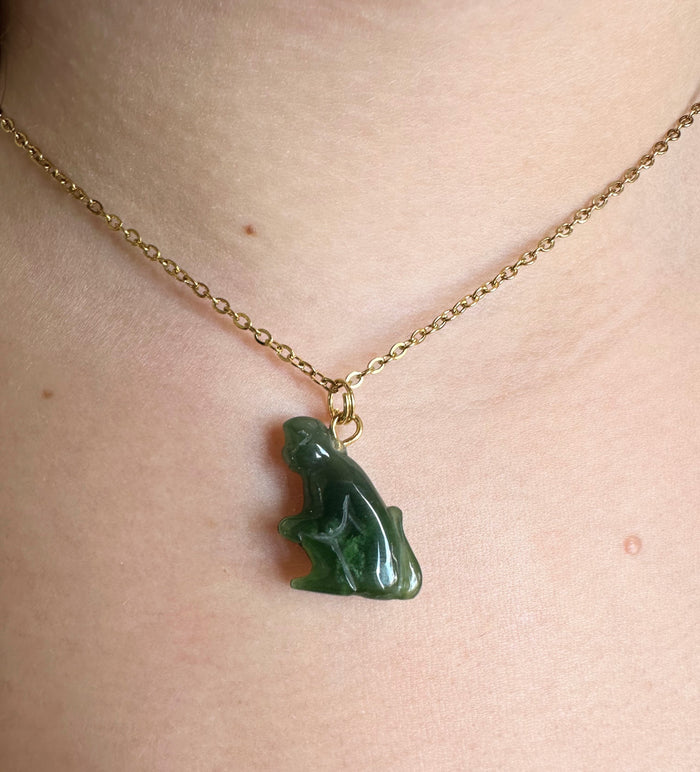 A close-up of a person's neck wearing a delicate gold chain necklace with a carved green jade monkey pendant.
