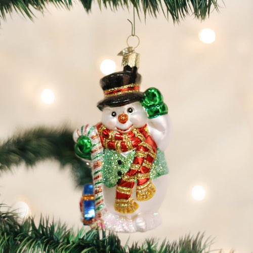 A festive glass snowman ornament with a black top hat, red and gold scarf, and green mittens hangs on a Christmas tree branch with blurred white lights in the background.