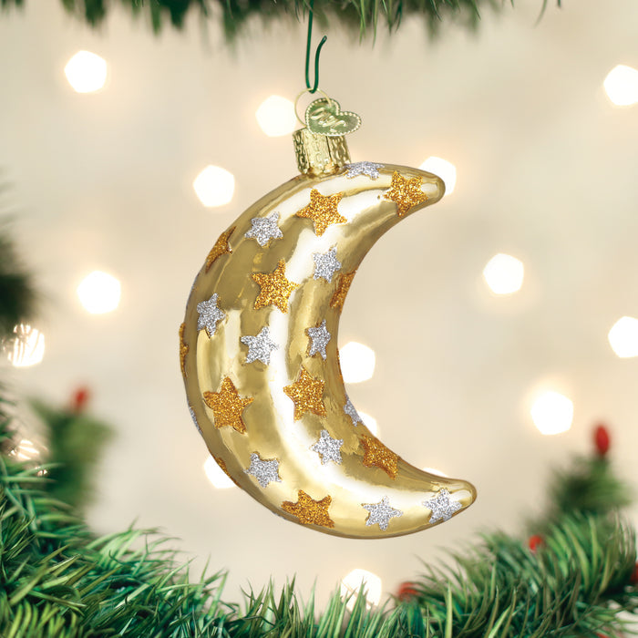 A gold crescent moon ornament decorated with gold and silver stars hangs on a Christmas tree, with blurred white lights in the background.