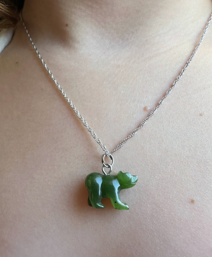 A close-up of a person's neck and chest wearing a silver chain necklace with a small carved green bear pendant.