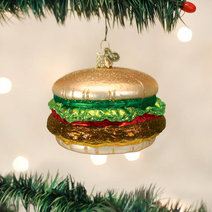 A shiny Christmas tree ornament shaped like a hamburger, featuring gold, green, red, and brown colors, hangs from a green tree branch with blurred lights in the background.