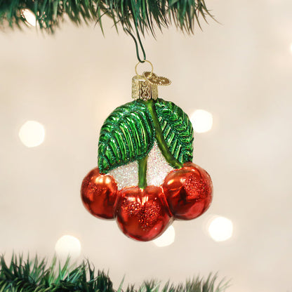 A shiny Christmas tree ornament shaped like three red cherries with two green leaves, hanging from a branch against a blurred background of white lights.