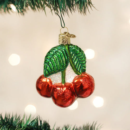 A shiny Christmas tree ornament shaped like three red cherries with two green leaves, hanging from a branch against a blurred background of white lights.