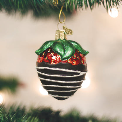 A Christmas tree ornament shaped like a chocolate-covered strawberry with green leaves and white drizzle, hanging from a branch with blurred lights in the background.