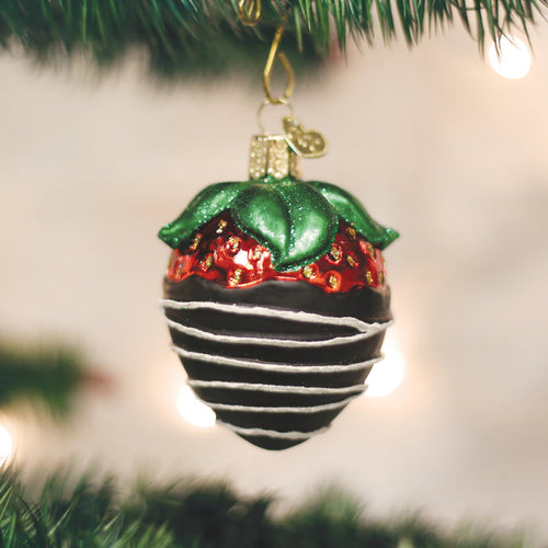 A Christmas tree ornament shaped like a chocolate-covered strawberry with green leaves and white drizzle, hanging from a branch with blurred lights in the background.