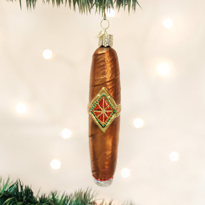 A shiny, brown, pickle-shaped Christmas ornament with a decorative green and red star design hangs from a green tree branch against a blurred background of white lights.