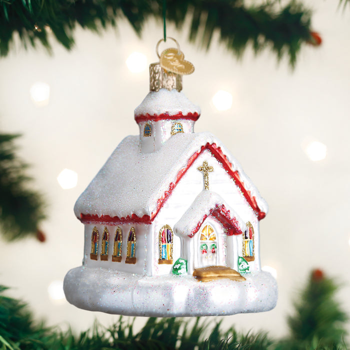 A glittery white Christmas church ornament with red trim hangs on a green tree branch, featuring golden windows, a gold cross, and snowy details against a blurred, light background.