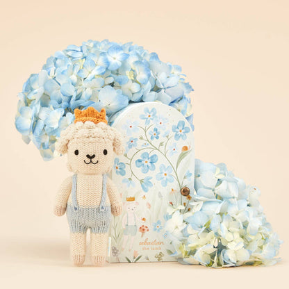 Knitted teddy bear with a crown, surrounded by blue flowers and a decorative pillow on a beige background
