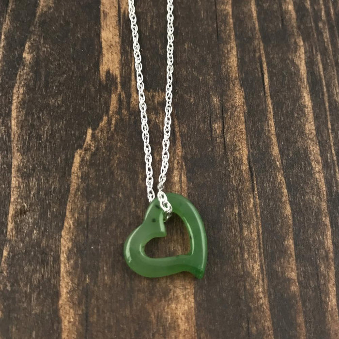 Green jade heart pendant with hollow center on a silver chain resting on dark wood surface