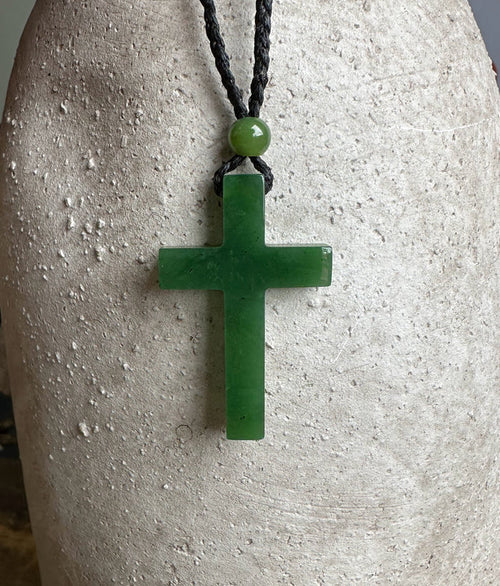 Dark green jade cross pendant on a black braided cord with a green bead above, displayed on a light textured surface