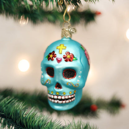 A blue sugar skull ornament decorated with colorful designs, including a yellow cross, hearts, and flowers, hangs on a Christmas tree with blurred lights in the background.