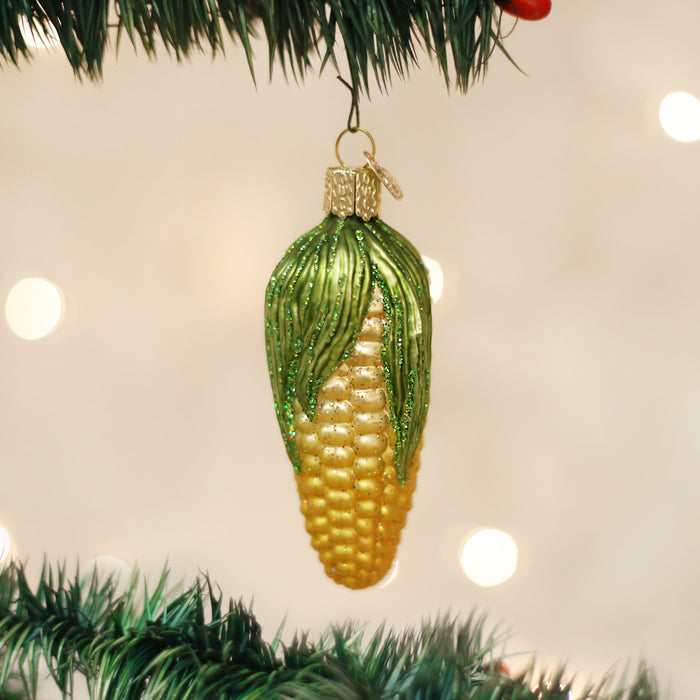 A Christmas tree ornament shaped like an ear of corn, with green husk details and a gold top, hanging from a tree branch with blurred lights in the background.