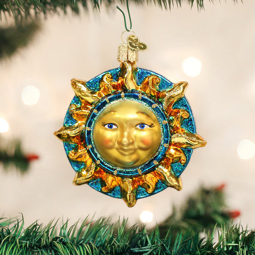 A glass Christmas tree ornament shaped like a sun with a smiling face, gold rays, and a blue glittery border hangs on a tree branch with blurred lights in the background.