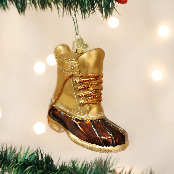 A Christmas tree ornament shaped like a golden and brown winter boot with gold laces hangs from a green tree branch, with soft white lights glowing in the background.