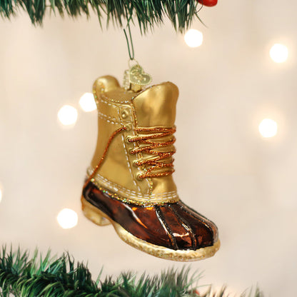 A Christmas tree ornament shaped like a golden and brown winter boot with gold laces hangs from a green tree branch, with soft white lights glowing in the background.