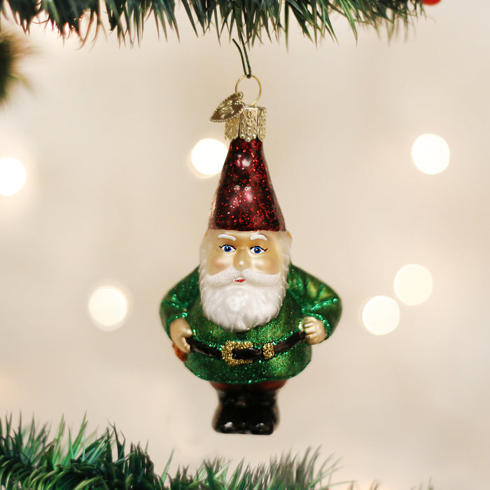 A glass Christmas ornament shaped like a bearded gnome in a green suit and red hat hangs from a decorated tree branch, with soft white lights blurred in the background.
