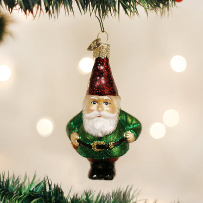 A glass Christmas ornament shaped like a bearded gnome in a green suit and red hat hangs from a decorated tree branch, with soft white lights blurred in the background.