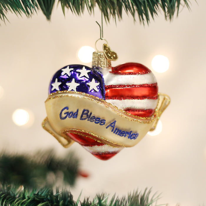 A heart-shaped Christmas ornament painted like the American flag with stars and stripes, featuring a gold ribbon that reads God Bless America, hanging on a Christmas tree.