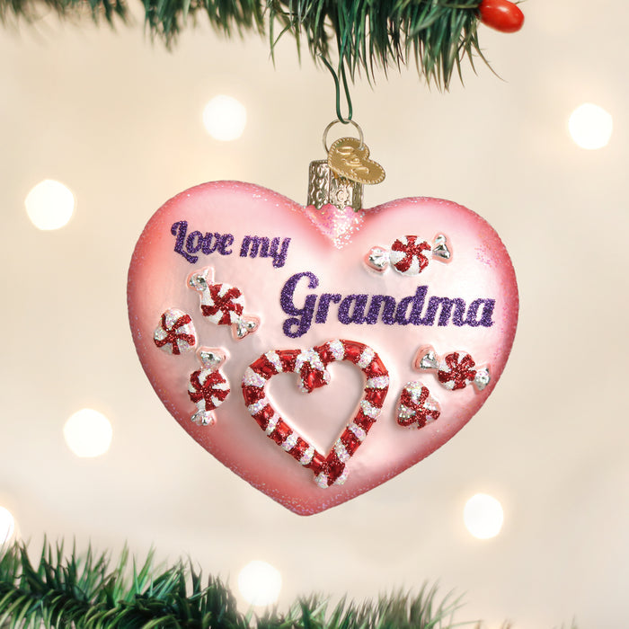 A pink heart-shaped Christmas ornament with â€œLove my Grandmaâ€_x009d_ written on it, decorated with red and white peppermint candy designs, hanging on a tree branch with blurred lights in the background.