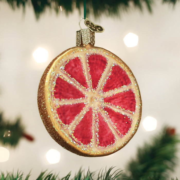 A Christmas tree ornament shaped and decorated like a glittery, red grapefruit slice hangs from a branch with soft lights in the background.