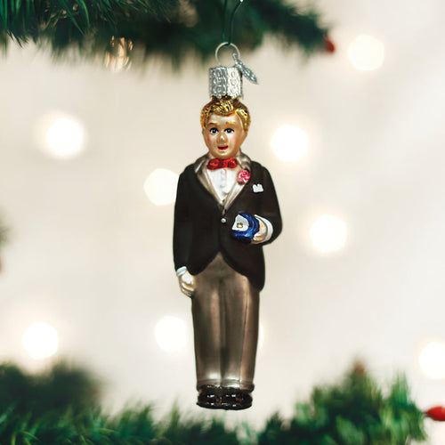 A Christmas tree ornament shaped like a man in a black tuxedo with a red bow tie and a boutonniere, holding a small blue and white object, hangs from a green tree branch with blurred lights in the background.