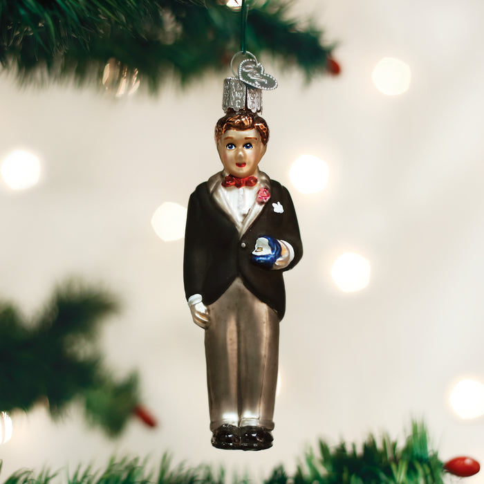 A Christmas tree ornament shaped like a man in a black tuxedo with a red bow tie, holding a small blue gift, hangs from a green branch with blurry white lights in the background.