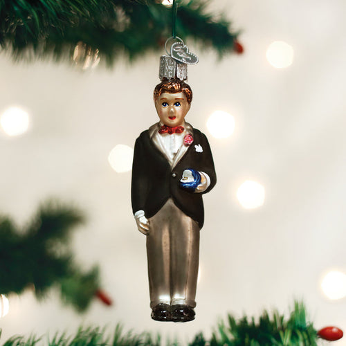 A Christmas tree ornament shaped like a man in a black tuxedo with a red bow tie, holding a small blue gift, hangs from a green branch with blurry white lights in the background.