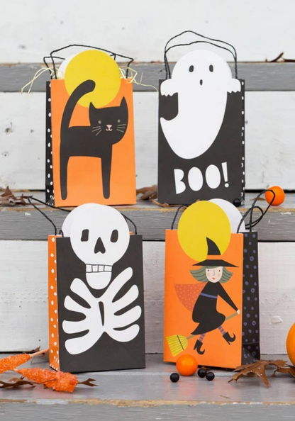 Four Halloween-themed paper treat bags arranged on rustic gray wooden steps. The bags are black and orange with white polka dots and feature illustrations of a black cat, a ghost with the word 'BOO!', a white skeleton, and a witch riding a broomstick.