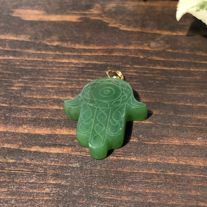 Green carved Hamsa hand pendant with swirling engravings and a gold bail on a wooden surface