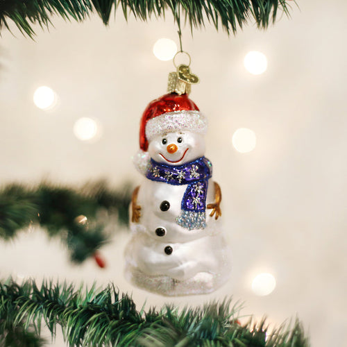 A glass snowman ornament with a red Santa hat and a blue scarf hangs on a Christmas tree. The background is blurred with warm white lights, creating a festive atmosphere.