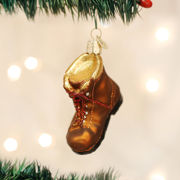 A Christmas tree ornament shaped like a brown boot with gold accents and red laces hangs from a green tree branch, with blurred white lights in the background.