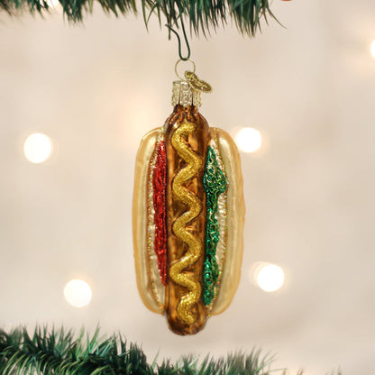 A Christmas tree ornament shaped like a hot dog with glittery toppings, hanging from a green branch with blurred lights in the background.