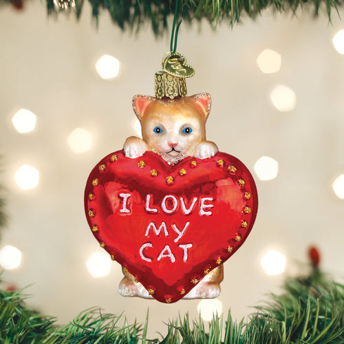 A Christmas tree ornament shaped like a tan cat holding a large red heart with the words I LOVE MY CAT written on it, hanging from a green branch with blurry lights in the background.