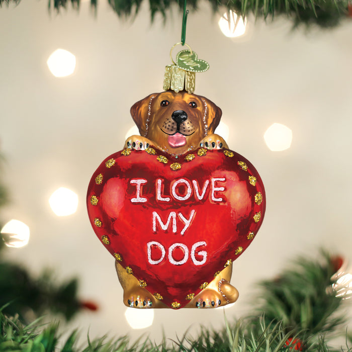 A Christmas tree ornament shaped like a brown dog holding a large red heart that says I LOVE MY DOG in white letters, hanging among green tree branches with blurred lights in the background.