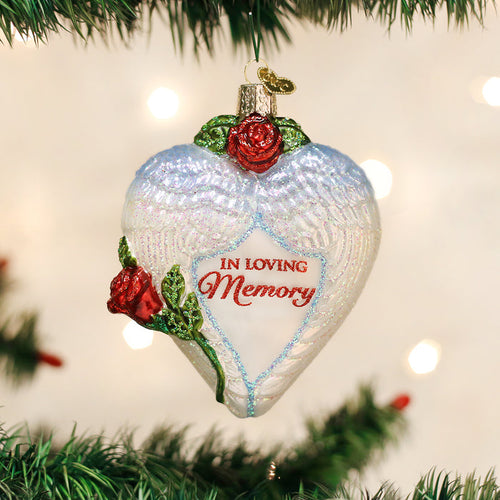 A heart-shaped ornament with angel wings, red roses, and the words In Loving Memory hangs on a Christmas tree with green branches and blurred lights in the background.