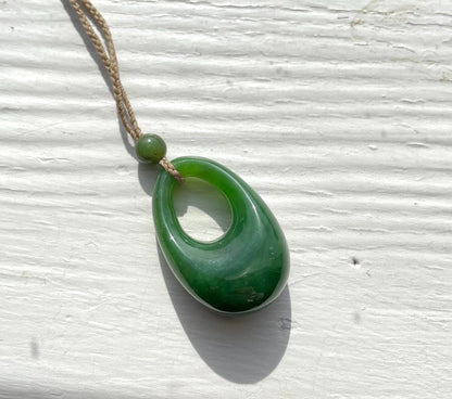 Green jade pendant with hole on a braided cord hanging against a pale textured wall